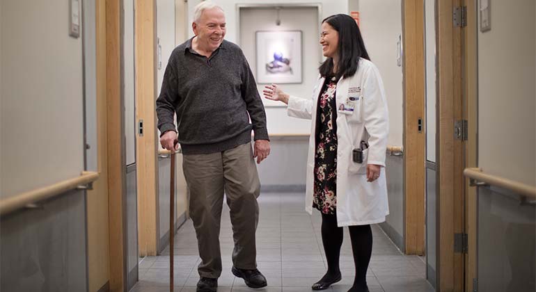 Male geriatric patient standing with cane in hallway alongside female doctor