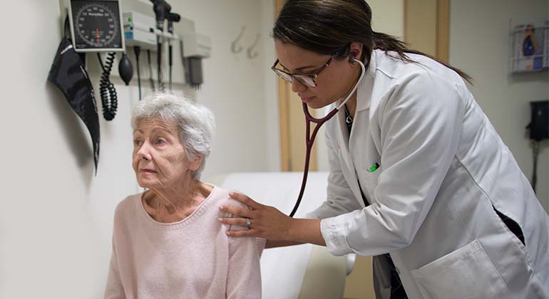 Female doctor using stethoscope on seated female geriatric patient in examination room