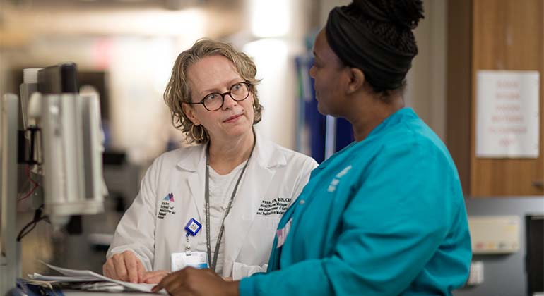 Female nurse manager and female nurse looking at each other while standing in front of table with papers on it