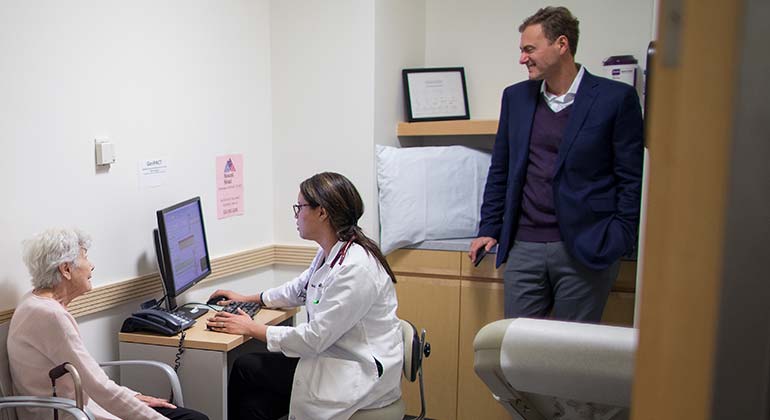 Female geriatric patient in examination room with seated medical professional and standing man