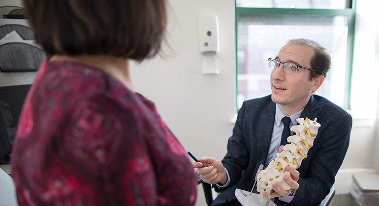 Seated doctor in a suit speaks with woman in a dress while holding a model spine