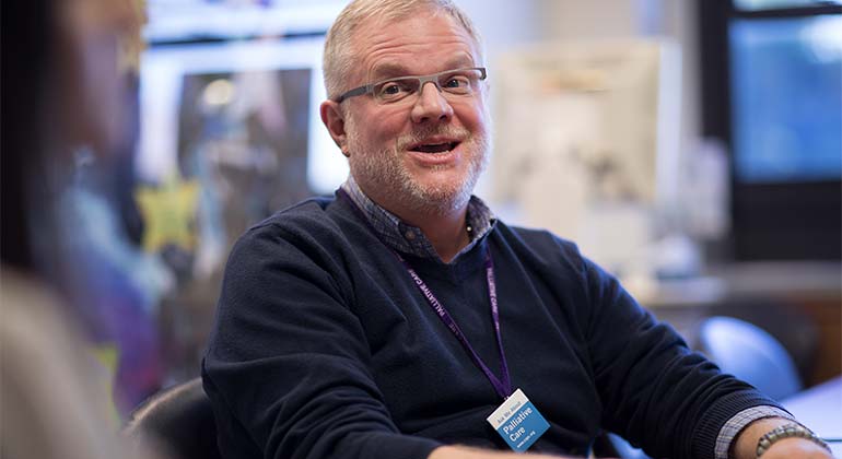 A man in glasses sitting at a table wearing a badge that says Ask me about palliative care