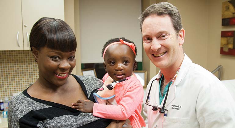 Dr. Peter Taub, pediatric plastic surgeon at Mount Sinai smiles and stands with toddler female patient and her mother after successful pediatric plastic surgery treatment.