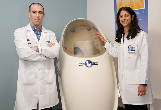 A photo of Reshmi Srinath, MD, and Avigdor Arad, PhD, RDN, CDE, at the Mount Sinai PhysioLab with one of their programs’ diagnostic tools, the Bod Pod, which measures the percentage of body fat and lean body mass.