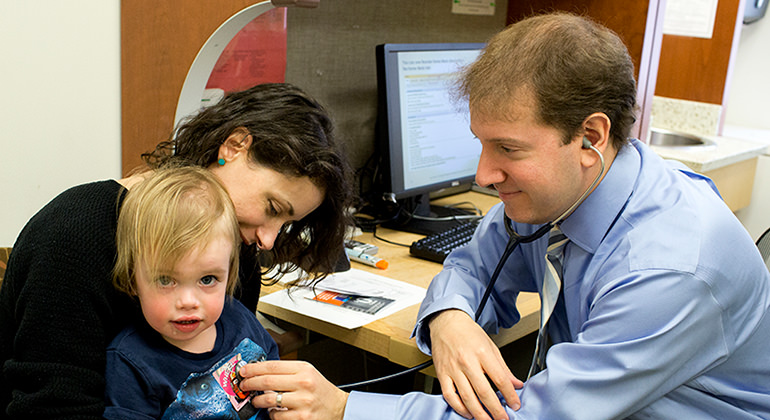 Allergist checking young boy sitting on mom’s lap with stethoscope