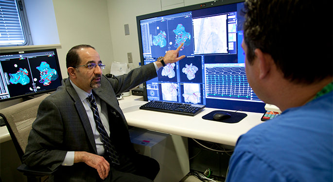 Seated man points at computer screen while talking to technician.