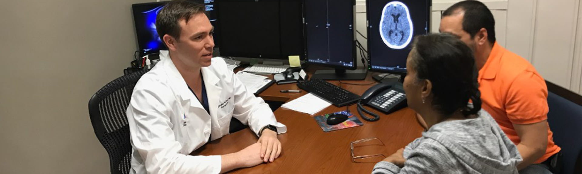 image of physician speaking to patients in his office, reviewing brain scan on computer screen 